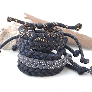 Puede incluir: Una pila de pulseras trenzadas de cuerda en varios tonos de negro, gris y azul. Algunas pulseras tienen patrones moteados en naranja, verde y blanco. Las pulseras son ajustables y tienen un tema náutico.