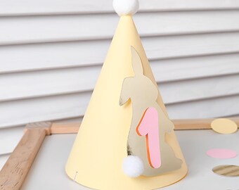 May include: Yellow paper party hat with a bunny silhouette and the number one in pink. The hat has a white pom-pom on top.