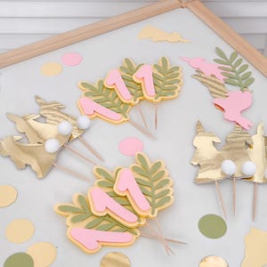 May include: A set of pink and yellow cake toppers featuring the number one, bunny silhouettes, and green leaves. The toppers are perfect for a first birthday party.