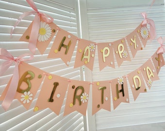 Op de afbeelding: Een roze en gouden banner met de woorden "Happy Birthday" in gouden letters en witte madeliefjes. De banner is aan elkaar geregen met roze lint.