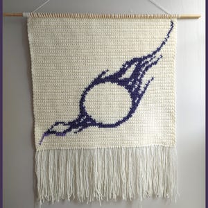 May include: Cream-colored, crocheted wall hanging with a purple comet design. The design features a central circle with a trailing tail. The bottom edge has a fringe. A wooden dowel and string are used for hanging.