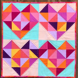 Pode incluir: Um edredão geométrico colorido com um padrão de coração repetido. O edredão apresenta uma paleta vibrante de rosas, roxos, laranjas e amarelos, com detalhes em azul claro e pêssego. O design é composto por triângulos e quadrados, com costuras visíveis.