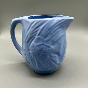 Puede incluir: Una jarra de cerámica azul claro con un diseño de pez estilizado. La jarra tiene una boca ancha y un asa.