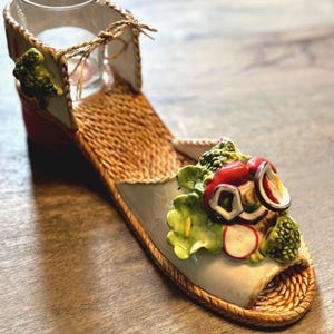 May include: A ceramic shoe shaped candle holder with a woven straw design. The shoe has a red, white, and green vegetable design on the toe.