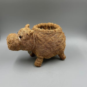 May include: A brown, woven hippo-shaped basket. The basket has a round opening and four short legs. The hippo has small, white eyes and a tag attached to its side. The basket is made of natural fibers and is suitable for home decor.