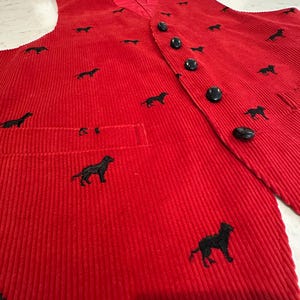 Puede incluir: Un chaleco de pana rojo con siluetas de perros bordadas en negro. El chaleco tiene cuello en V, un bolsillo y botones negros. La tela tiene una textura acanalada vertical.