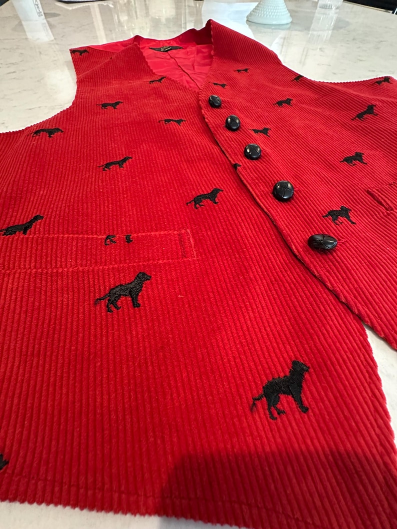 Puede incluir: Un chaleco de pana rojo con siluetas de perros bordadas en negro. El chaleco tiene cuello en V, un bolsillo y botones negros. La tela tiene una textura acanalada vertical.