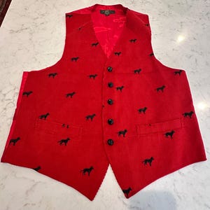 Puede incluir: Un chaleco de pana rojo con un estampado de siluetas de perros negros. El chaleco tiene cuello en V, botones negros y dos bolsillos delanteros. El forro es rojo.