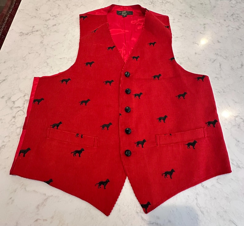 Puede incluir: Un chaleco de pana rojo con un estampado de siluetas de perros negros. El chaleco tiene cuello en V, botones negros y dos bolsillos delanteros. El forro es rojo.