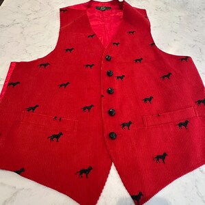 Puede incluir: Un chaleco de pana rojo con un estampado de perro negro. El chaleco tiene cuello en V, botones negros y dos bolsillos delanteros. El forro interior es rojo.