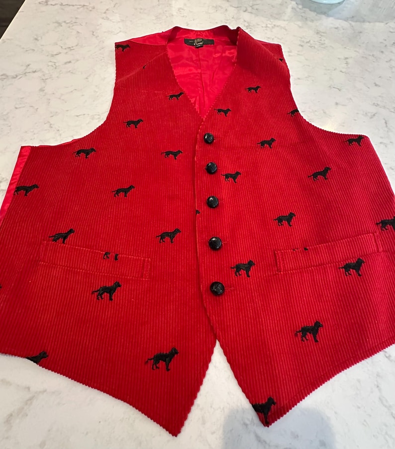 Puede incluir: Un chaleco de pana rojo con un estampado de perro negro. El chaleco tiene cuello en V, botones negros y dos bolsillos delanteros. El forro interior es rojo.