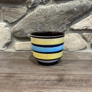 May include: A ceramic planter with a yellow and blue striped design. The planter has a black rim and base. The interior is dark brown. The planter is sitting on a wooden surface.