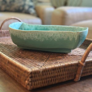 May include: A rectangular, turquoise ceramic bowl with a textured, white rim sits on a woven rattan tray with handles. The bowl has a curved shape and is likely used for serving food or as a decorative piece. The tray is brown.