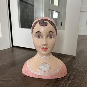 May include: A decorative bust of a woman with a pink and white headscarf and dress. The bust has painted facial features, including brown eyes and red lips. A small plaque on the bust reads "Chapeau Pour Rosette".