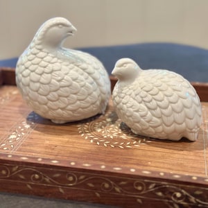 May include: Two off-white ceramic quail figurines with textured feather details sit on a decorative wooden tray. The tray has an inlaid floral design and a dark brown border. The quail are positioned at different angles, suggesting a natural pose.