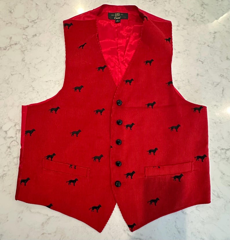 Puede incluir: Un chaleco de pana rojo con un estampado de siluetas de perros negros. El chaleco tiene cuello en V, forro de sat&eacute;n rojo y botones negros. Cuenta con dos bolsillos delanteros y un dise&ntilde;o cl&aacute;sico.