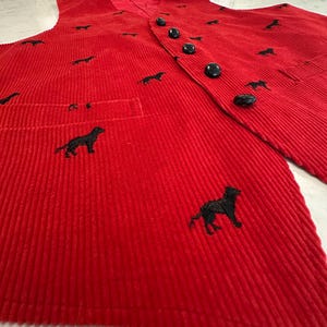 Puede incluir: Un chaleco de pana rojo con bordados de perros negros. El chaleco tiene una parte delantera abotonada, un bolsillo y un forro de sat&eacute;n rojo. Los botones negros combinan con el bordado.