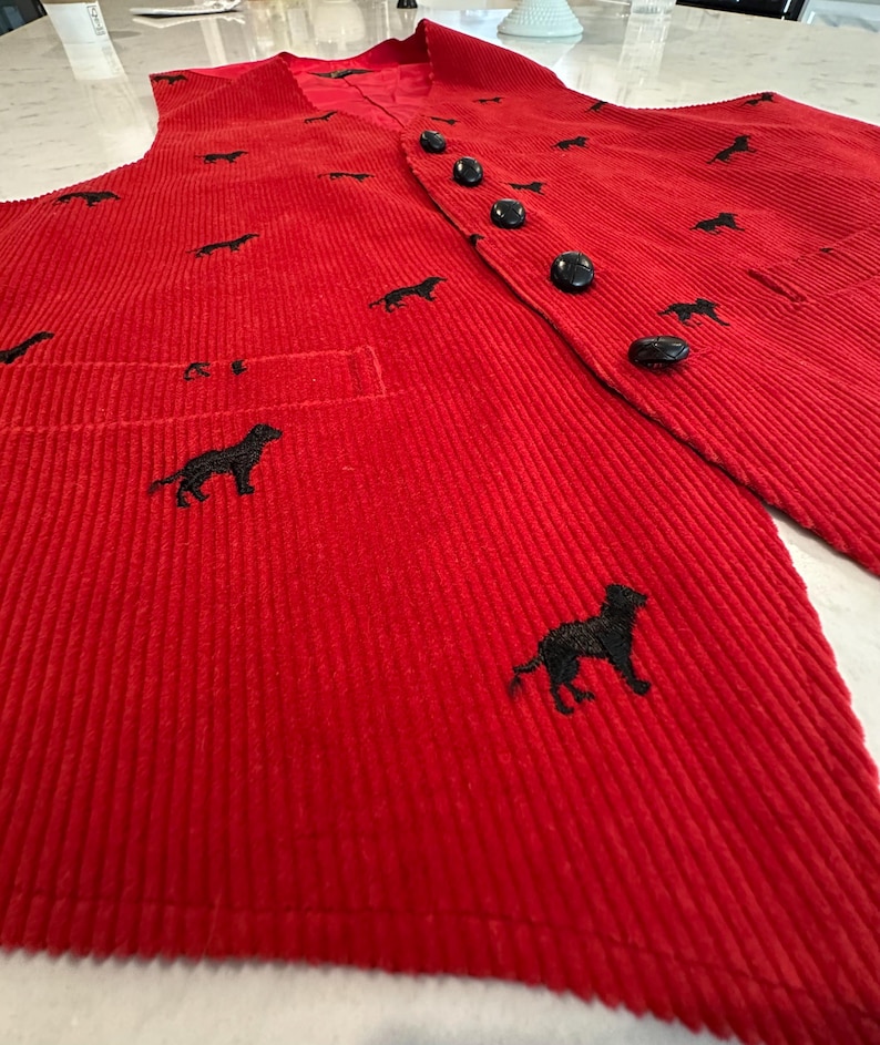 Puede incluir: Un chaleco de pana rojo con bordados de perros negros. El chaleco tiene una parte delantera abotonada, un bolsillo y un forro de sat&eacute;n rojo. Los botones negros combinan con el bordado.
