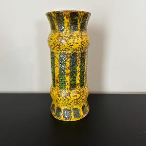May include: A cylindrical ceramic vase with a yellow base color and a textured surface. The vase features vertical stripes of green and brown, with a flared top and base. The vase is approximately 10 inches tall.