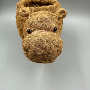 May include: A handcrafted, brown woven basket shaped like a hippopotamus head. The basket features dark, round eyes and a detailed snout. The open top provides space for storage or planting. The basket is made of natural fibers.