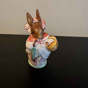 May include: A ceramic figurine of a rabbit wearing a red and white striped bonnet and a white dress with a red collar. The rabbit is holding a yellow basket and a green umbrella. The base of the figurine is striped with purple and pink.