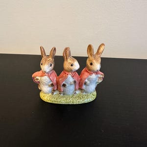May include: A ceramic figurine featuring three brown rabbits standing side by side on a green textured base. Each rabbit has white fur on its chest and wears a small red cape. The rabbits have pointed ears, dark eyes, and are facing forward.