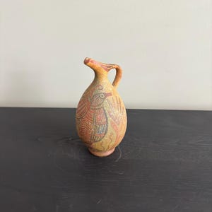 May include: A small, hand-painted ceramic jug with a spout and handle. The jug is decorated with a red, yellow, and black geometric pattern.