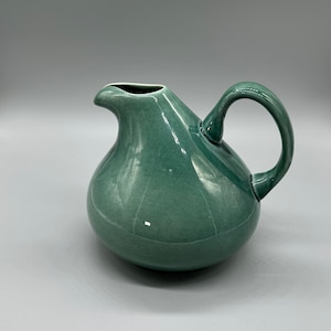 May include: A green ceramic pitcher with a rounded body and a curved handle. The pitcher has a smooth, glossy finish.