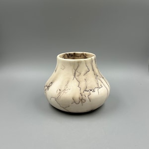 May include: A small, white ceramic vase with a rounded bottom and a wide opening. The vase has a unique marbled pattern in shades of brown.