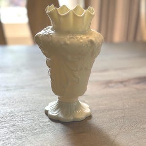 May include: A white porcelain vase with a ruffled top and intricate floral designs. The vase has a classic, elegant style.