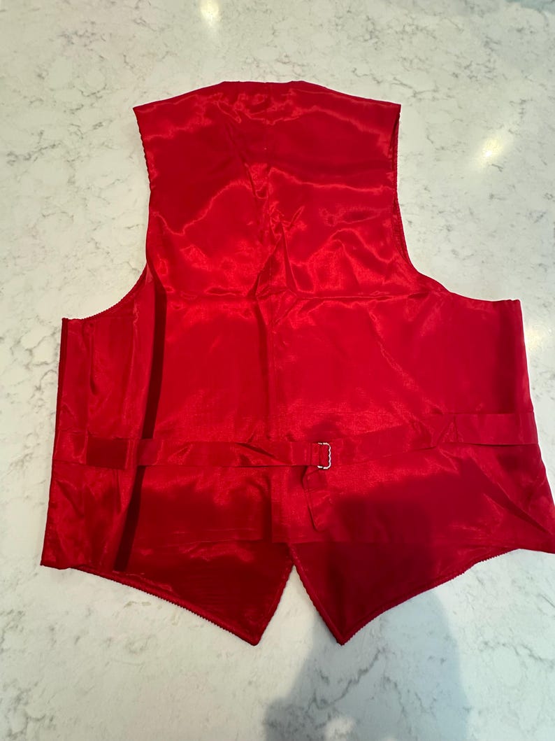 Puede incluir: Un chaleco rojo brillante hecho de un material brillante similar al sat&eacute;n. La parte trasera del chaleco es visible, mostrando una correa ajustable con una hebilla plateada. El chaleco tiene una forma cl&aacute;sica y a medida.