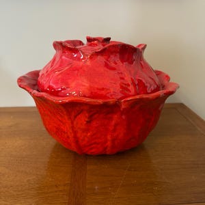 May include: A bright red ceramic lidded bowl shaped like a head of cabbage. The bowl features a textured, glossy surface with realistic leaf-like folds and edges. The lid is domed and mimics the layered appearance of vegetable leaves.