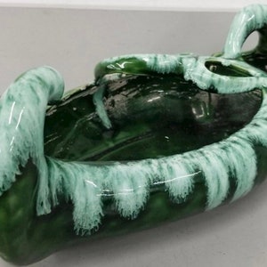 May include: A green ceramic planter with white glaze, featuring a unique, organic shape with two handles. The planter has a smooth, glossy finish and a textured, flowing design.