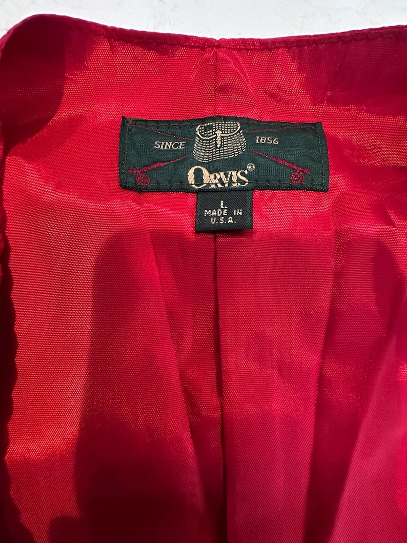 Puede incluir: Primer plano del interior de una prenda roja con una etiqueta verde. La etiqueta dice "Orvis" e incluye el texto "Since 1856" y "Made in U.S.A.". La prenda es de un rojo s&oacute;lido y vibrante.