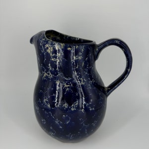 May include: A blue ceramic pitcher with a white marbled design. The pitcher has a rounded body and a curved handle.