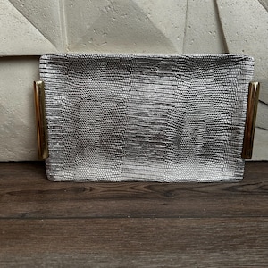 May include: A rectangular silver tray featuring a textured, reptilian-inspired pattern across its surface. The tray is equipped with two smooth, polished gold-toned metal handles attached to the left and right sides, providing a contrast in color and finish.