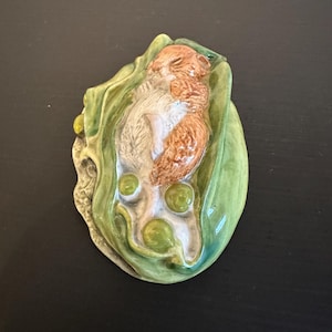 May include: A ceramic decorative piece featuring a small, brown and white hamster sleeping inside a green pea pod. The pod is shaped like a curved leaf with three round, green peas visible near the bottom. The surface has a glossy finish.