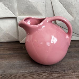 May include: A round, glossy pink ceramic pitcher featuring a wide, bulbous body, a curved handle on the right side, and a prominent pouring spout. The surface of the pitcher is smooth and reflective, showcasing a solid, uniform pastel pink color.