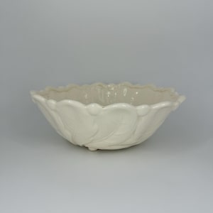 May include: A white ceramic bowl with a leaf design. The bowl has a scalloped edge and three small feet.