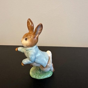 May include: A ceramic figurine of a brown rabbit standing on a green base. The rabbit has long ears and is wearing a light blue jacket. The figure is depicted in a dynamic pose with one arm extended forward, featuring a glossy, smooth finish.