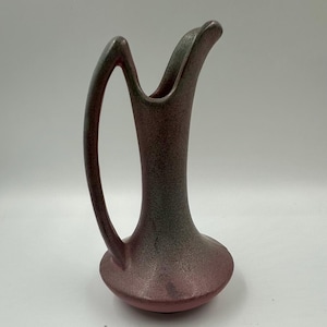 May include: A vintage, small pitcher with a handle. The pitcher is a muted pink with a speckled texture and a green-gray rim. The handle is a curved, open design. The pitcher has a rounded base and a slender neck.