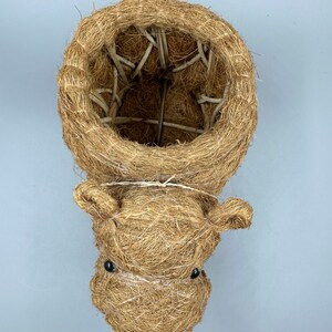 May include: A handcrafted, brown, woven fiber hanging planter shaped like a hippo's head. The planter has a round opening and small, black bead eyes. The hippo's head is attached to the planter with a thin, white string.