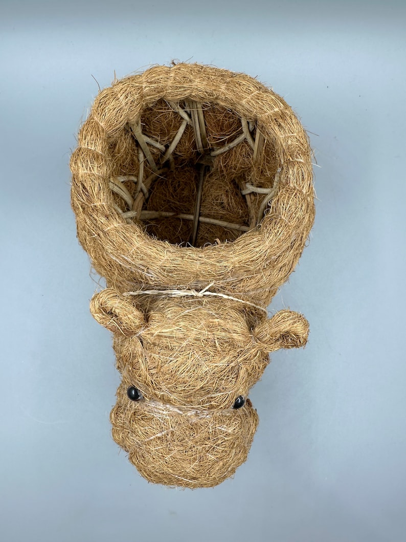 May include: A handcrafted, brown, woven fiber hanging planter shaped like a hippo's head. The planter has a round opening and small, black bead eyes. The hippo's head is attached to the planter with a thin, white string.