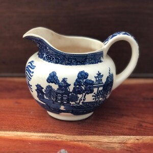 May include: A small, white ceramic creamer with a blue Willow pattern. The creamer has a spout, a curved handle, and a round body. The blue pattern depicts a landscape with buildings and trees. The creamer sits on a wooden surface.