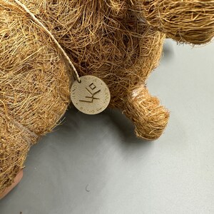 May include: Close-up of a brown, woven decorative object, possibly a figurine or sculpture. A small, round tag is attached with a string. The tag has text and a symbol. The object is made of natural fibers.