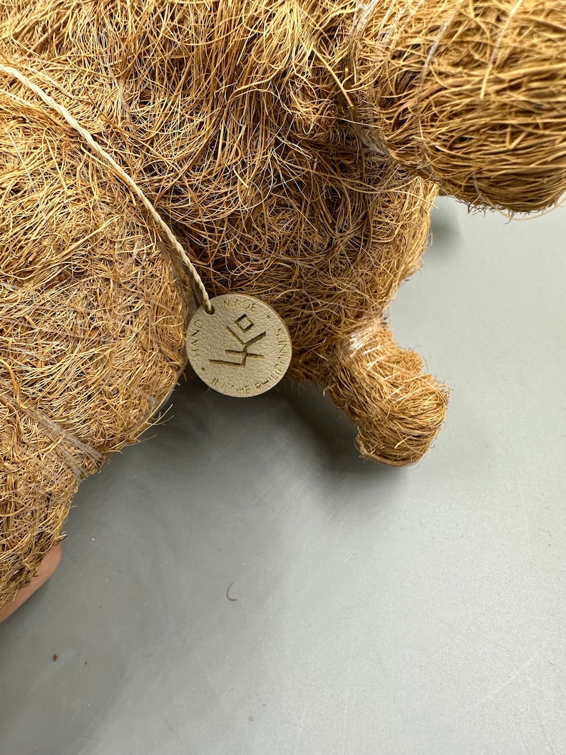 May include: Close-up of a brown, woven decorative object, possibly a figurine or sculpture. A small, round tag is attached with a string. The tag has text and a symbol. The object is made of natural fibers.