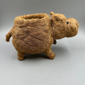 May include: A brown, hippo-shaped planter made from woven natural fibers. The planter has a wide opening for plants and small legs. A small bell hangs from a string around the hippo's neck. The hippo planter is a decorative item for home decor.
