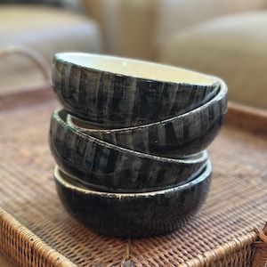 May include: A stack of four ceramic bowls with a black and gray striped pattern. The bowls have a cream-colored interior and are stacked on a woven wicker tray. The background is blurred, suggesting a home setting.