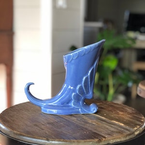 May include: A blue ceramic vase shaped like a shoe with a pointed toe and a curled heel. The vase is sitting on a wooden stool.