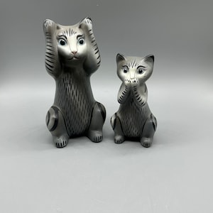 May include: Two ceramic cat figurines, one larger than the other, both painted in shades of gray with black stripes. The larger cat is covering its ears with its paws, while the smaller cat is covering its mouth with its paws.